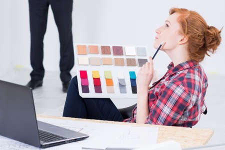 Young and red haired woman designer sitting thoughtful at a desk with a laptop on, color picker in...