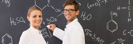 Students in lab clothes writing chemical formulas on blackboard