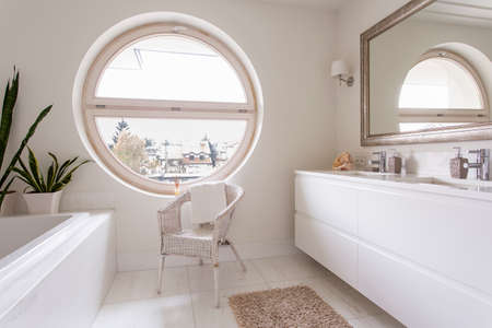 Modern, white bathroom with large round window, mirror, bathtub and chairの写真素材