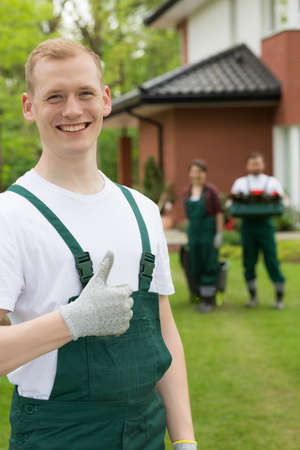 Young smiling gardener standing with the thumb up and two oder gardeners at the backgroundの写真素材