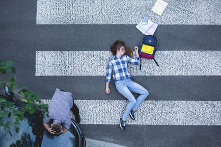 Female student hit by a car, lying on the road next to her school suppliesの写真素材