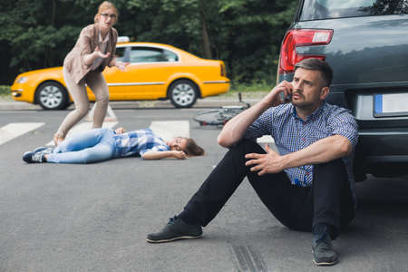 Shot of a shocked man sitting next to his car and calling for help while unconscious victim of the car crash is lying in the backgroundの写真素材