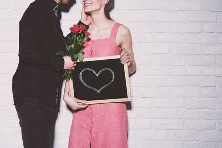 Young woman holding a small blackboard with heart on it and receiving roses from her boyfriendの写真素材