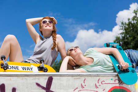 Two girls lying on a vert ramp with skateboards and looking at the blue skyの写真素材