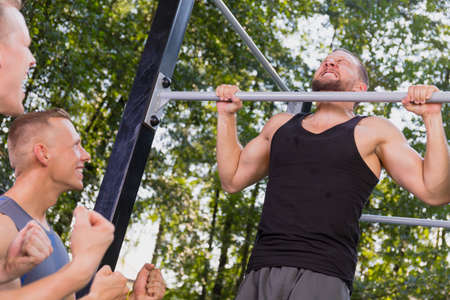 Handsome man doing pull-ups during outdoors training with friends