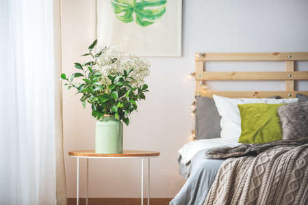 Bunch of flowers on small table next to bed in cozy bright bedroomの写真素材