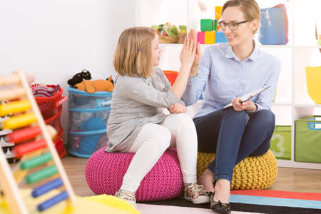 Blond girl and  smiling woman sitting on colorful poufs and giving each other high fiveの写真素材