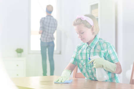 Girl wiping wooden table and her mum cleaning the windowsの写真素材