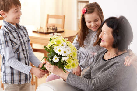 Happy grandchildren giving a bunch of flowers to their grandma sitting on a sofaの写真素材