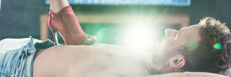 Handsome man laying on a bed with his lover's foot in red high heel touching his body