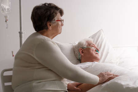 Sad senior wife supporting dying ill husband in hospital bed