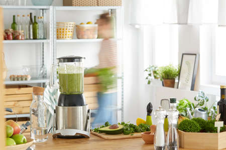 Wooden kitchen table with green vegetables and blender with smoothieの写真素材
