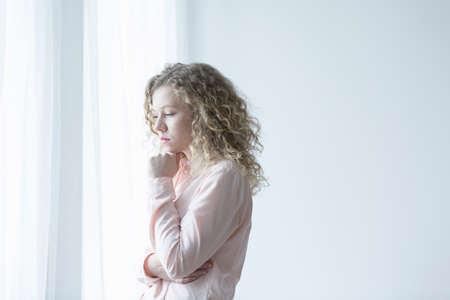 Young depressed woman standing next to the windowの写真素材