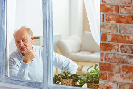 Sad wondering senior man looking out the window in apartmentの写真素材