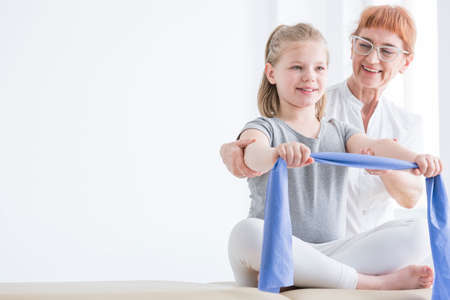 Female physiotherapist supervising little girl during stretching the rubber tape