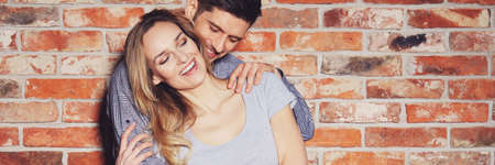 Happy couple in casual clothes hugging and joking against brick wallの写真素材