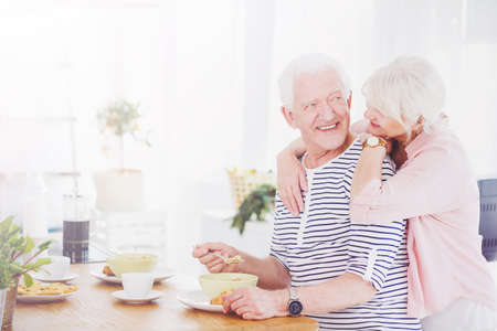 Senior man eating breakfast looking at his wife hugging himの写真素材