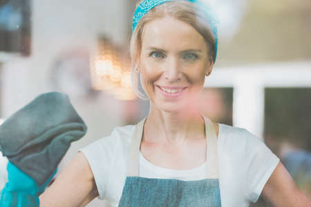 Happy housekeeper with blue band on head is cleaning the window with grey towelの写真素材