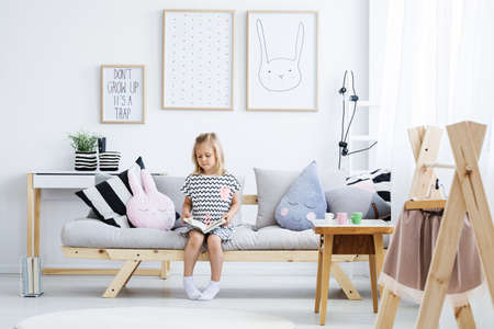 Cute young girl reading book on stylish sofa in white roomの写真素材