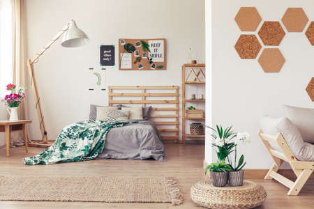 Wooden bed and couch in botanic open interior with cork honeycomb wall, jute rug, rattan pouf and green plantsの写真素材