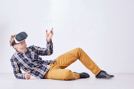 Blonde man in casual outfit lies on floor in white room with VR glasses on head
