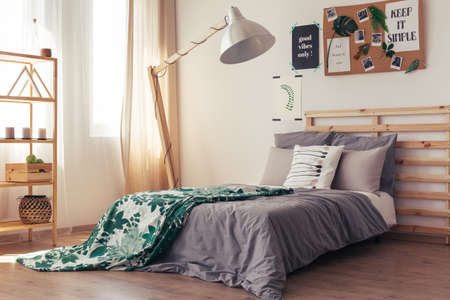 Contemporary natural bedroom with white wall, cork pin board, lamp, wooden furniture and botanic duvet coverの写真素材