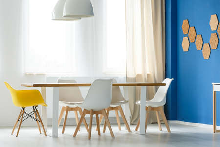Multifunctional scandinavian home interior with communal table, white and yellow chairs, blue wall,...