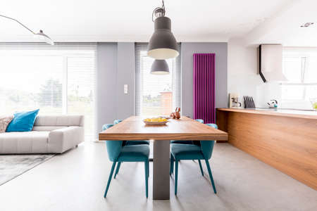 Open plan apartment of family living-space with wooden kitchen countertop, purple radiator, communal table with turquoise chairs and spacious living room with beige couchの写真素材