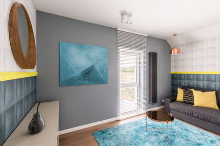 Trendy sophisticated interior with copper coffee table, abstract painting, turquoise rug, mirror, gray couch and yellow detailsの写真素材