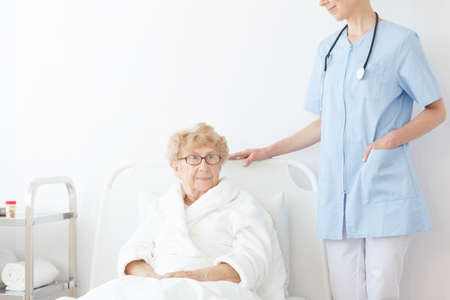 Young doctor in blue uniform is standing by the bed of a sick elderly woman in glassesの写真素材
