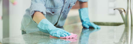 Woman in jean shirt, and rubber gloves cleaning a kitchen countertop with a pink dishrag