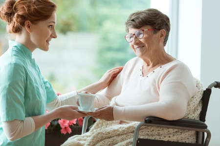 Disabled elder woman sitting in the wheelchair with blanket and nurse giving her a cup of tea