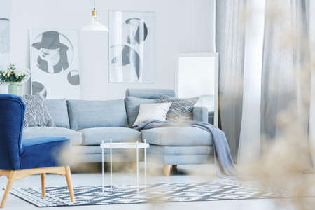 Blue armchair in modern living room with patterned pillows on grey sofa against wall with fashionable postersの写真素材