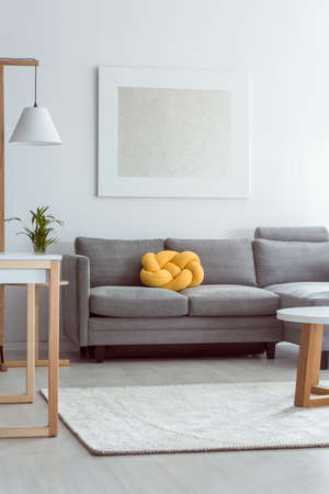 Yellow knot pillow on grey sofa in cozy living room with white carpet on floor and simple painting on wallの写真素材