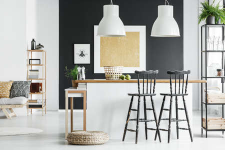 White lamps above kitchen countertop with bar stools and braided pouf on wall in room with gold...