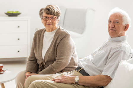 Couple of elder people sitting together on white coach next to coffee table
