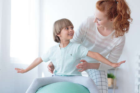 Red haired physiotherapist supporting boy with scoliosis doing correction exercise on mint ballの写真素材
