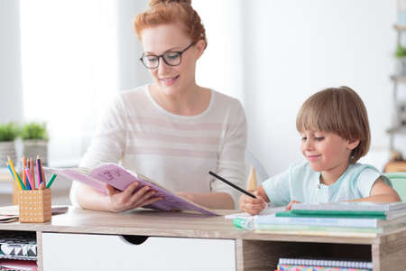Math teacher doing exercises from workbook with young happy boy while sitting at desk in classroom