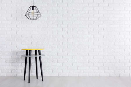 Designer lamp above creative table with yellow stool with black legs against white brick wallの写真素材
