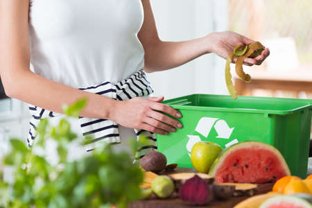 Woman recycling organic kitchen waste by composting in green container during preparation of mealの写真素材