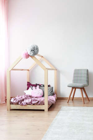 Grey patterned chair next to handmade bed with cute pillows and pink overlay in bright kids bedroom...