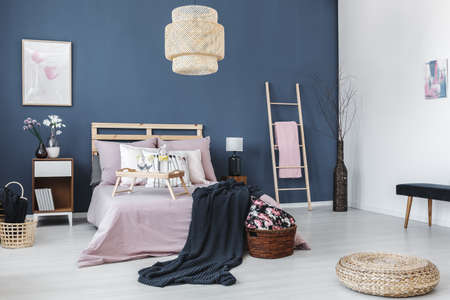 Bedside cabinet with black lamp between ladder with pink towel and king-size bed against blue wallの写真素材