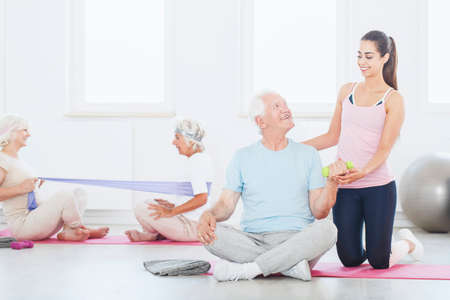 Happy elderly person sitting with crossed legs on pink mat and lifting a dumbbell with trainer supporting himの写真素材
