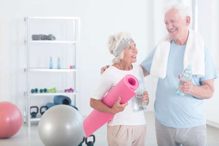 Grandmother holding pink fitness mat and smiling to grandfather before yoga classes in fitness studioの写真素材