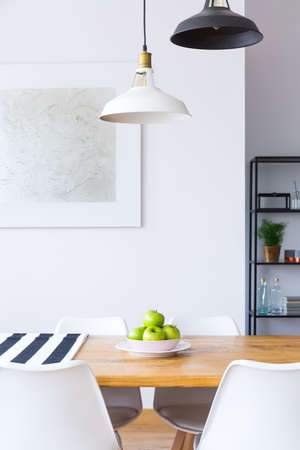 Lamps above dining table with apples and white chairs in contrast color room with painting