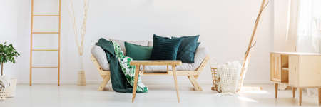Beige knit blanket in basket next to sofa with moss green pillows in living room with ladder and...