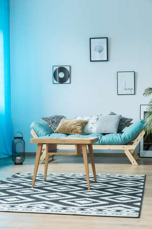 Posters on wall above blue sofa with pillows in cozy living room with table on geometric carpetの写真素材