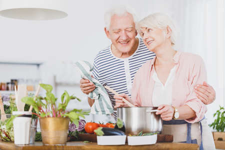 Grandfather is watching grandma who mixes vegetable soup in a pot during cooking dinnerの写真素材
