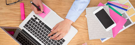 High angle of a person making notes while using laptop on desk, preparing business plan for start-up