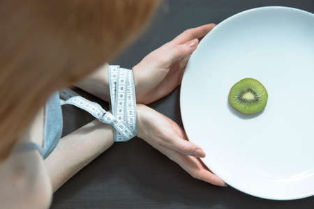 Woman with an eating disorder, and her hands bound by tape-measure limiting her dinner to a kiwiの写真素材
